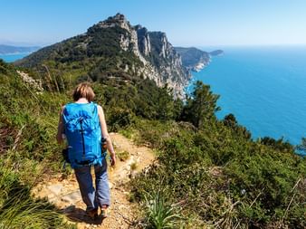 Wanderer mit blauem Rucksack auf Bergpfad in Cinque Terre mit Blick auf türkisfarbenes Mittelmeer und dramatische Felsenküste.