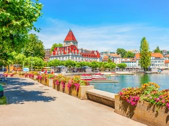 Schloss Ouchy mit rotem Turm und weißer Fassade am Ufer des Genfersees in Lausanne. Promenade mit bunten Blumen und Booten im Hafen.