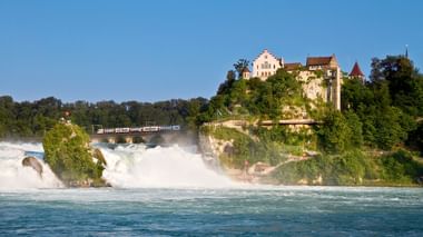 Rheinfall-Wasserfall mit weißem kaskadierendem Wasser und Nebel. Das historische Schloss Laufen thront auf grünen Klippen darüber.