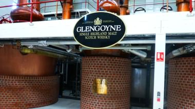 Innenraum der Glengoyne-Destillerie mit großen Kupferbrennblasen und ziegelverkleideten Gärtanks mit dem Glengoyne Single Highland Malt Schild.