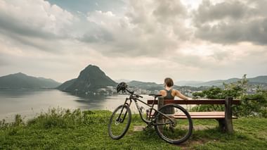 Eine Frau sitzt auf einer Bank mit Sicht auf den Lago di Lugano und den San Salvatore.