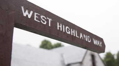 Brauner Holzwegweiser mit weißen Buchstaben 'WEST HIGHLAND WAY' vor verschwommenem Hintergrund mit Bäumen und Gebäuden.