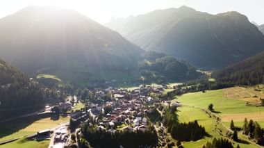 Luftaufnahme des Dorfes Bergün in einem Tal, umgeben von bewaldeten Bergen, grünen Wiesen und traditionellen Alpengebäuden.