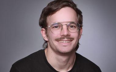 Professionelles Portrait von Rafael Kälin mit Brille und dunklem Shirt, der warm in die Kamera lächelt vor grauem Hintergrund.
