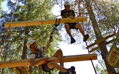 Zwei Kinder mit Schutzhelmen und Gurten bewältigen Holzplattformen und Balken in einem Waldseilgarten zwischen hohen Bäumen.