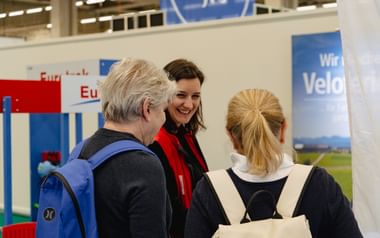 Eurotrek-Mitarbeiterin in rot-schwarzer Weste berät zwei Besucher am Messestand in Bern. Ausstellungsdisplays im Hintergrund sichtbar.