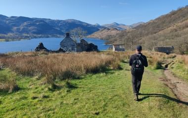 Wanderer mit Rucksack auf Grasweg zu Steinruinen neben schottischem See, mit Bergen im Hintergrund unter blauem Himmel.