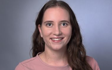 Professionelles Porträt von Juliana Grella mit langen braunen Haaren, rosa Oberteil und kleinen Ohrringen, lächelnd vor grauem Hintergrund.