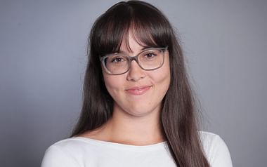 Porträt von Anja Kostidis mit langen dunklen Haaren, Brille und weißem Oberteil vor grauem Hintergrund.