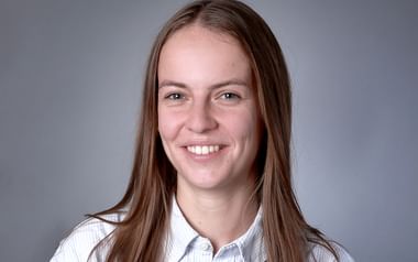 Professionelles Porträt von Livia Weber mit langen braunen Haaren, weißem Hemd mit Kragen und warmem Lächeln vor grauem Hintergrund.