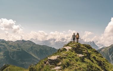 Zwei Wanderer stehen auf einem schmalen grasbewachsenen Berggrat der Via Glaralpina in Glarus, mit dramatischen Alpengipfeln und Wolken im Hintergrund.