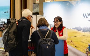 Mitarbeiterin in roter Weste berät Besucher am Eurotrek-Reisestand. Display zeigt malerische Radroute mit grünen Wiesen und Bergen.