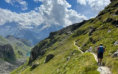 Reto wandert mit blauem Rucksack auf Bergpfad der Haute Route zum Lac de Louvie, umgeben von grünen Alpenhängen und schneebedeckten Gipfeln.
