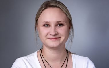 Professionelles Porträt von Anna-Maria Beirid mit blonden Haaren, weißem Oberteil und schwarzer Kordel-Halskette, lächelnd vor grauem Hintergrund.