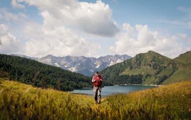 Wanderer in roter Jacke geht durch gelbgrüne Wiese zum Ritomsee im Tessin, mit bewaldeten Bergen und schneebedeckten Gipfeln unter bewölktem Himmel.