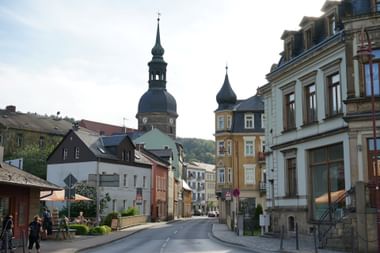 Straßenansicht in Bad Schandau mit bunten historischen Gebäuden und einer Kirche mit dunklem Kuppelturm. Fußgänger gehen entlang der Straße.