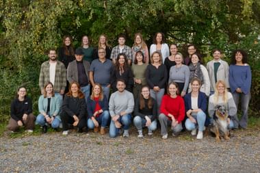 Teamfoto mit Kollegen und Hund im Freien Gruppe von etwa 25 Personen in drei Reihen für ein Teamfoto im Freien arrangiert, mit grünem Laubhintergrund und einem Hund in der ersten Reihe.