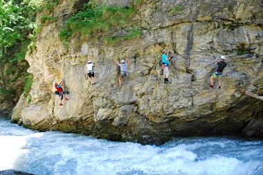 Six climbers with helmets and harnesses scaling a tall limestone rock face above rushing white water in Tirol, Austria.