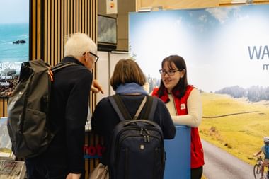 Mitarbeiterin in roter Weste berät Besucher am Eurotrek-Reisestand. Display zeigt malerische Radroute mit grünen Wiesen und Bergen.