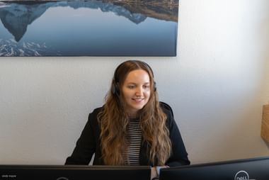 Frau mit Headset und langen braunen Haaren lächelt am Computer im Büro. Bergsee-Foto hängt an weißer Wand hinter Schreibtisch.