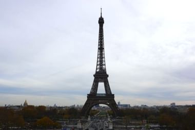 Der Eiffelturm erhebt sich über der Pariser Skyline unter bewölktem Himmel. Bäume mit Herbstlaub rahmen die Basis, Stadtgebäude sind in der Ferne sichtbar.