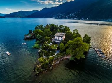 Luftaufnahme der Brissago Inseln im Lago Maggiore mit weißer Villa umgeben von üppiger grüner Vegetation, Boote in der Nähe, Berge im Hintergrund.