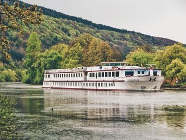 Weißes Flusskreuzfahrtschiff MS Normandie fährt auf ruhigem Wasser mit bewaldeten Hügeln im Hintergrund. Das mehrstöckige Schiff spiegelt sich im Wasser.
