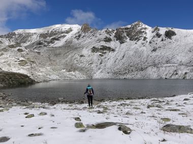 Wanderin mit Rucksack am verschneiten Ufer des Bergsees Meidsee, vor schneebedeckten Gipfeln unter blauem Himmel.