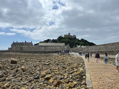 Steinerner Damm zum St Michael's Mount in Cornwall mit Wanderern auf felsigem Strand bei Ebbe unter bewölktem Himmel.