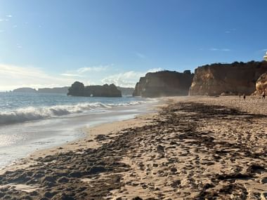 Sandstrand am Praia do Amado mit Fußspuren im Sand, anrollenden Wellen und dramatischen Felsformationen entlang der Algarve-Küste.