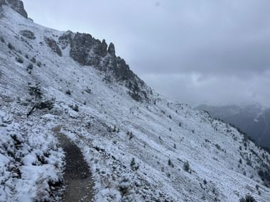 Verschneiter Bergweg an steilem Hang mit Felsgipfeln und vereinzelten Bäumen. Bewölkter Himmel mit Nebel im Tal auf dem Alpenpässeweg.
