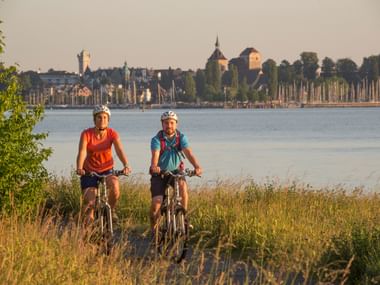 Zwei Radfahrer mit Helmen auf Fahrrädern am Bodensee, mit einer historischen Stadt mit Kirchtürmen und Hafen sichtbar über das Wasser.