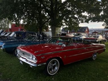 Rotes Oldtimer-Cabrio im Vordergrund einer Oldtimer-Show in Trosa, mit mehreren Oldtimern auf Rasen unter großen Bäumen.