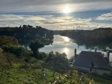 Malerischer Blick auf einen ruhigen Fluss in Tréguier, Bretagne, mit bewaldeten Ufern und Häusern. Sonne durchbricht Wolken und spiegelt sich im Wasser.