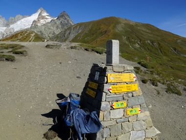 Vorne ein blauer Rucksack der an einem Grenzstein angelehnt ist, welcher auch Gleich als Wegweiser genutzt wird. Im Hintergrund die Spitze des mit Schnee bedeckten Mont-Blanc