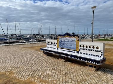 Weiße Fliesenbank mit blauem Azulejo-Panel und goldenem Rahmen im Hafen von Faro. Marina mit Segelbooten und bewölktem Himmel im Hintergrund.