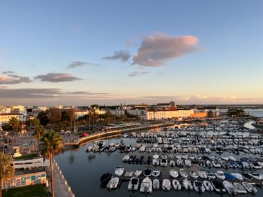Luftaufnahme des Faro-Hafens voller Boote bei Sonnenuntergang. Historische Stadtgebäude und Uferpromenade unter pastellfarbenem Himmel.