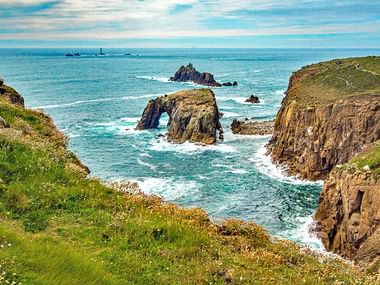 Dramatische Küstenansicht in Cornwall mit natürlichem Felsbogen im türkisfarbenen Wasser, steilen, grün bewachsenen Klippen und Schiffen am Horizont.