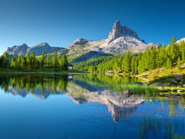 Lago Federa in the Italian Dolomites with crystal clear water perfectly reflecting dramatic rocky peaks and green coniferous forest under blue sky.