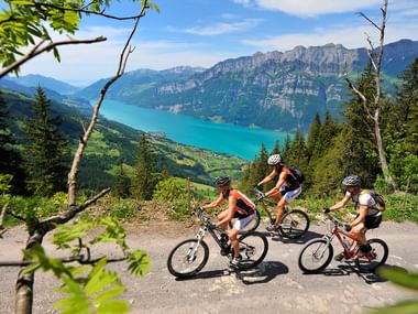 Drei Mountainbikerinen trampem den Aufstieg vor den Bergen und einem Türkisblauen See im Tal durch.