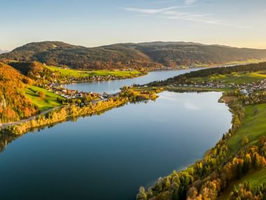 Luftaufnahme eines Sees im Schweizer Jura, umgeben von bewaldeten Hügeln und grünen Wiesen mit kleinen Dörfern zur goldenen Stunde.