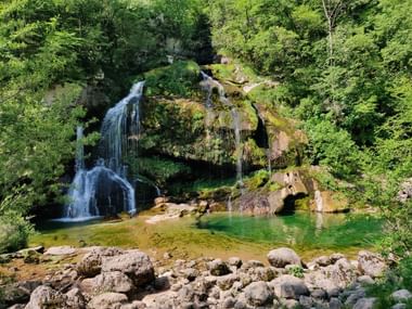 Breiter Wasserfall fällt über mit Moos bewachsenen Steine - umgeben von Bäumen.