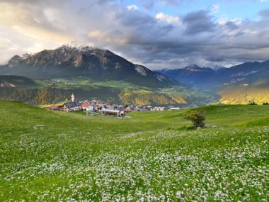 Dorf Stierva im Schweizer Alpental Das Dorf Stierva eingebettet in ein grünes Schweizer Alpental mit schneebedeckten Bergen im Hintergrund und einer blumenreichen Wiese im Vordergrund.