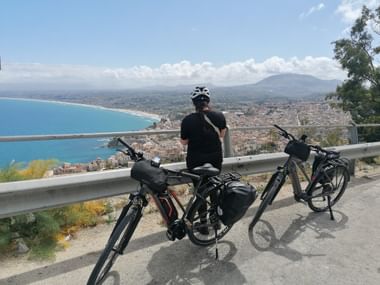 Radfahrer ruht an Leitplanke mit zwei Reiserädern und Blick auf Castellammare Bucht und Küstenstadt in Sizilien mit Bergen im Hintergrund.