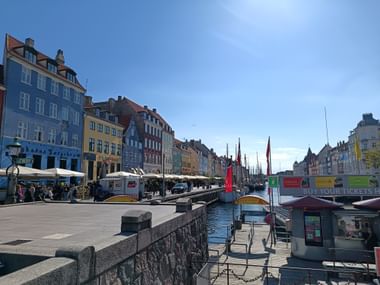 Sonnige Ansicht von Kopenhagens Nyhavn-Hafen mit bunten historischen Gebäuden, Kanalbooten, Straßencafés und Touristen entlang der Uferpromenade.