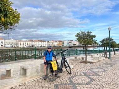 Radfahrer mit blauer Weste und Helm steht mit Tourenrad am Fluss Gilão in Tavira. Historische Gebäude sind über das Wasser sichtbar.