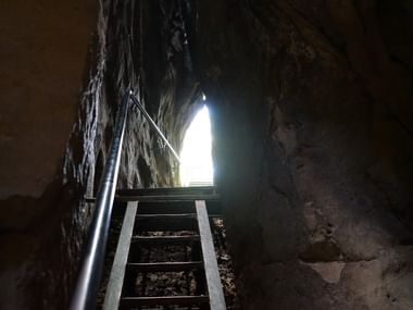 Metalltreppe mit Geländer führt durch engen Felsdurchgang nach oben zu heller Öffnung. Steinwände rahmen den steilen Aufstieg am Malerweg.