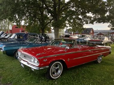 Rotes Oldtimer-Cabrio im Vordergrund einer Oldtimer-Show in Trosa, mit blauen und anderen Klassikern auf Rasen unter großen Bäumen.