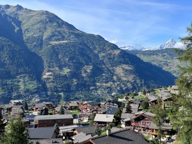 Alpendorf Grächen an einem Berghang mit traditionellen Chalets, umgeben von bewaldeten Hängen und schneebedeckten Gipfeln unter blauem Himmel.