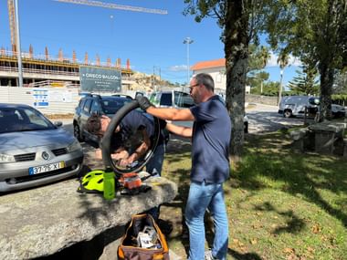 Mann in blauem Hemd repariert Fahrradreifen im Freien in Ponte de Lima. Fahrradrad, Helm und Reparaturwerkzeug auf Gras neben parkenden Autos.